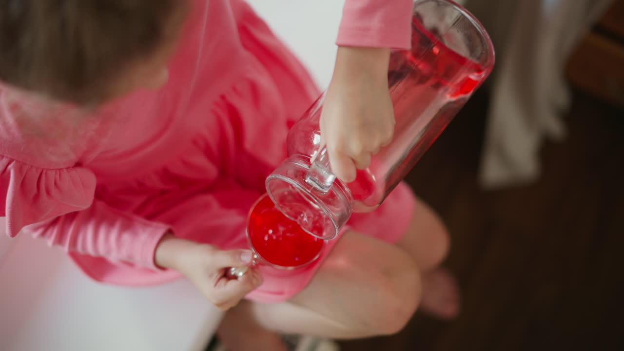 Aerial view of school girl seated by window lifting glass jar to pour red juice into cup, spilled drink soaking pink dress and sill, small hands gripping handle and curious expression on child