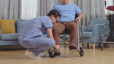 asian male nurse helping a patient in wheelchair putting feet on footrest after a physical therapy at home