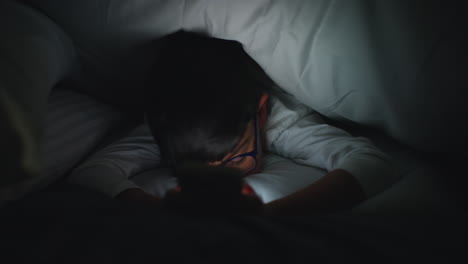 primer plano de un niño en el dormitorio en casa usando el teléfono móvil para enviar mensajes de texto bajo cubiertas o edredón por la noche 7