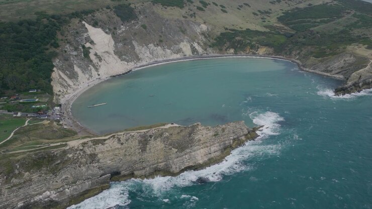 Veduta aerea ravvicinata di Lulworth Cove nel Dorset che mostra la caratteristica baia curva, l'acqua turchese, le scogliere calcaree e le onde che si infrangono lungo la Jurassic Coast