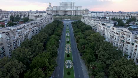 Impresionante-Vista-Aérea-En-Cámara-Lenta-Del-Palacio-Del-Parlamento-En-Bucarest,-Rumania-Al-Atardecer,-Con-Cielo-Naranja-Y-Fuentes-De-Agua-Danzantes