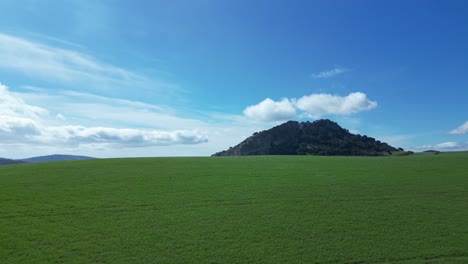 Flying-with-a-drone-over-a-green-meadow-with-some-spectacular-mountains-in-the-background-with-a-blue-sky-with-white-clouds