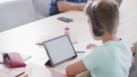 Fröhlicher-Kaukasischer-Junge,-Der-Am-Tisch-Sitzt-Und-Ein-Tablet-Mit-Kopierraum-Benutzt,-Zeitlupe