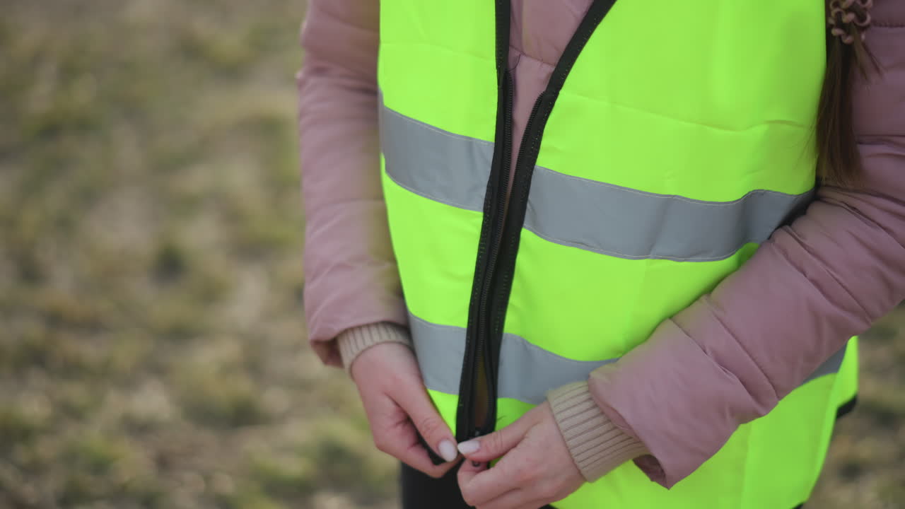 Close-up of woman in pink winter jacket and beige sweater fastening zipper on bright yellow reflective safety vest outdoors, preparing for work, volunteering, or visibility task in cool weather conditions