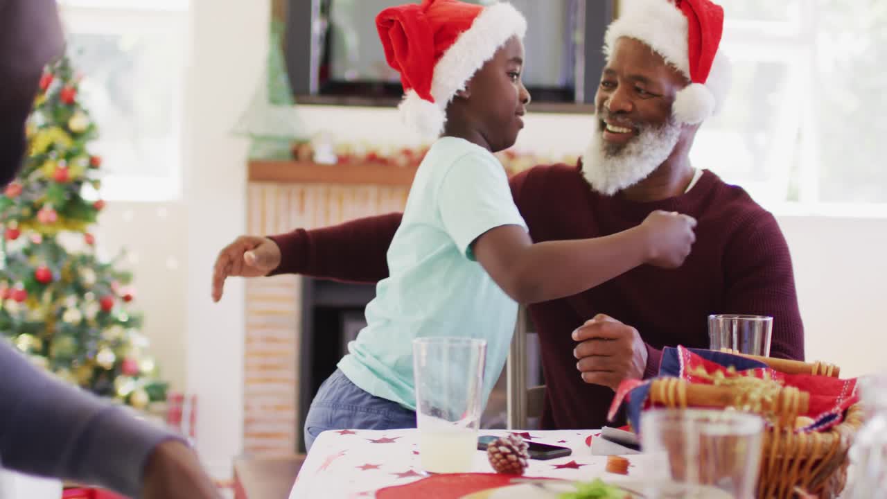 niño afroamericano en sombrero de santa abrazando a su abuelo mientras estaba sentado en la mesa de comedor almorzando