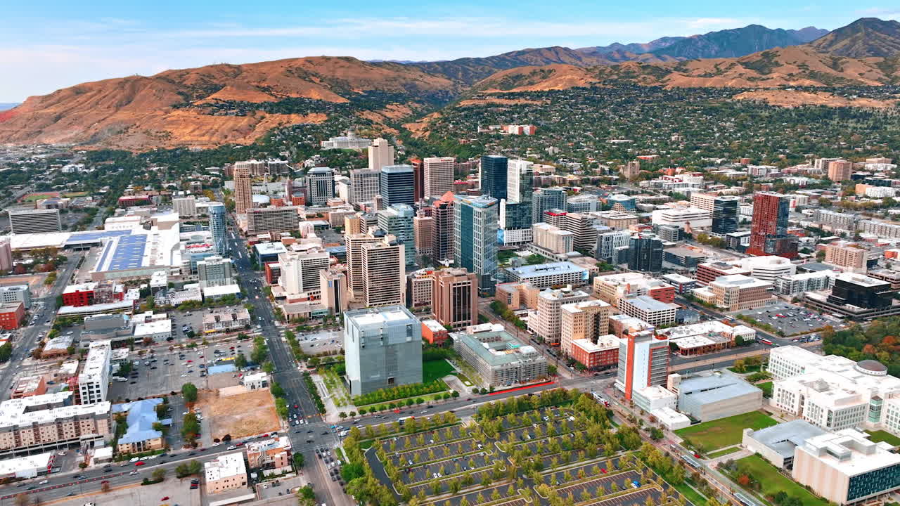 Distancing from a green car park with planted trees. Numerous vehicles ride by the roads. Rocks covered with residential area at backdrop. Salt Lake City, Utah, USA. Aerial view