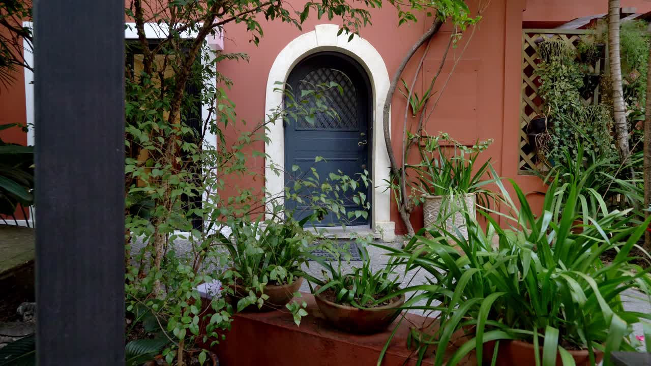 A charming arched doorway with a dark blue door is framed by lush greenery and potted plants, set against a warm terracotta wall with a white trim.