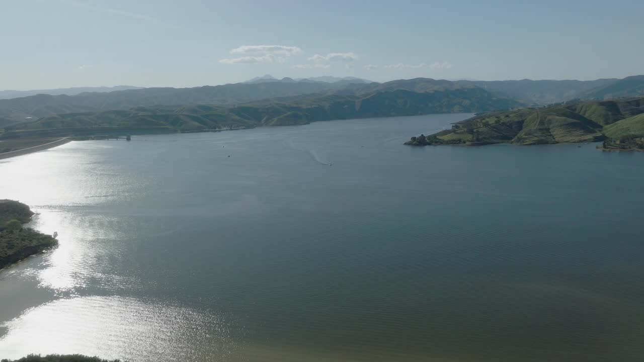 Aerial shot of Castaic Lake in Santa Clarita, California with green hillsides and several small watercraft moving over the water and the Castaic dam just showing on the far left.