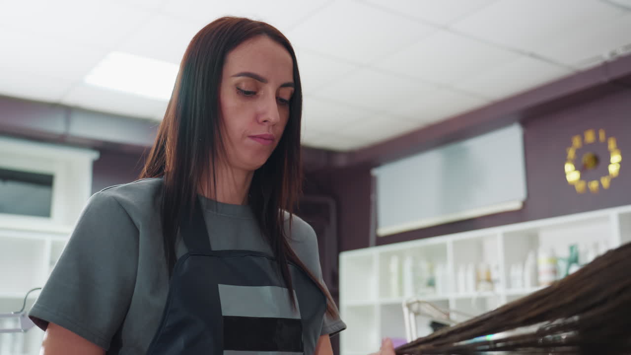 Estilista caucásica concentrada en una sección de pelo con expresión de concentración, manos trabajando suavemente entre los mechones, estantes de salón y suministros en un fondo suave, habilidad profesional a la vista