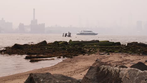 view of manhattan covered in smoke from wildfires seen from beach on the east river, waves crashing on sandy beach and rocks in the foreground and ferry crossing frame