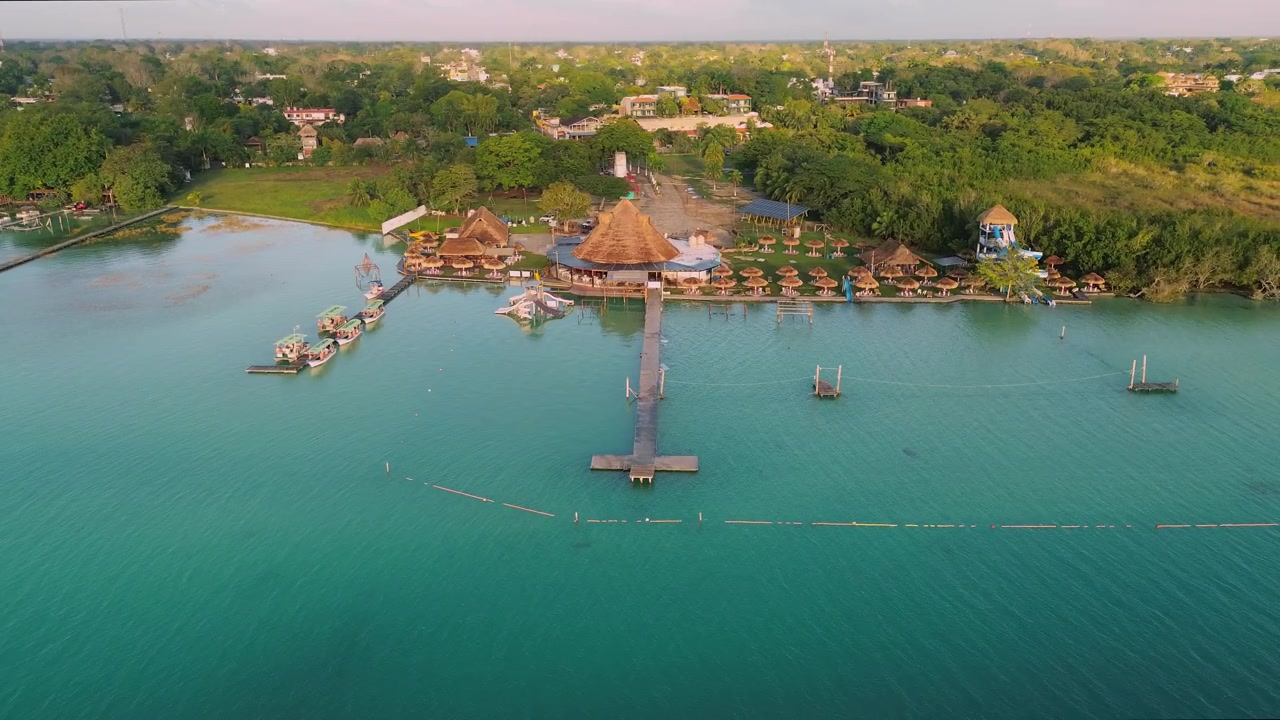 Riprese con drone con panoramica a sinistra su un resort sul lago all'alba, che mostrano palapas con tetto di paglia, una torre di un parco acquatico, moli galleggianti e barche turistiche sulle acque turchesi della Laguna di Bacalar, Messico
