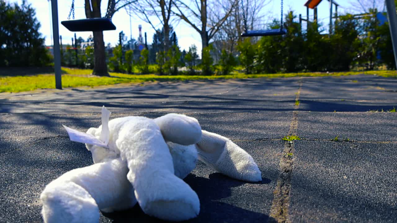 Coniglio di peluche bianco dimenticato che giace sul terreno asfaltato in un parco giochi per bambini deserto con altalene che si muovono sullo sfondo, simboleggiando solitudine, perdita e infanzia perduta.