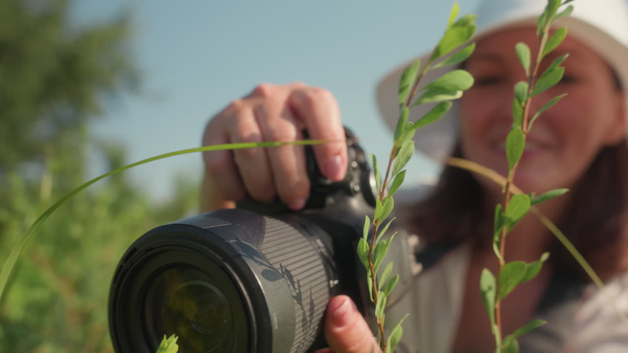 Close up of smiling woman in white sun hat squatting in grassy field using camera to film tall green plants under bright daylight with focus on capturing beauty of wild nature