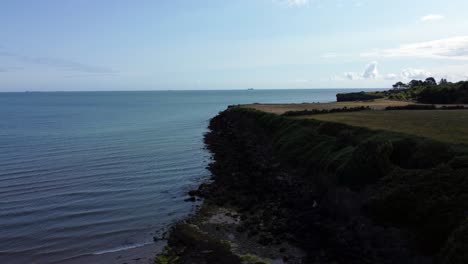 Traeth-Lligwy-idyllic-rocky-coast-shoreline-aerial-view-pull-back-dolly-across-green-pasture-on-rocky-cliffs-edge