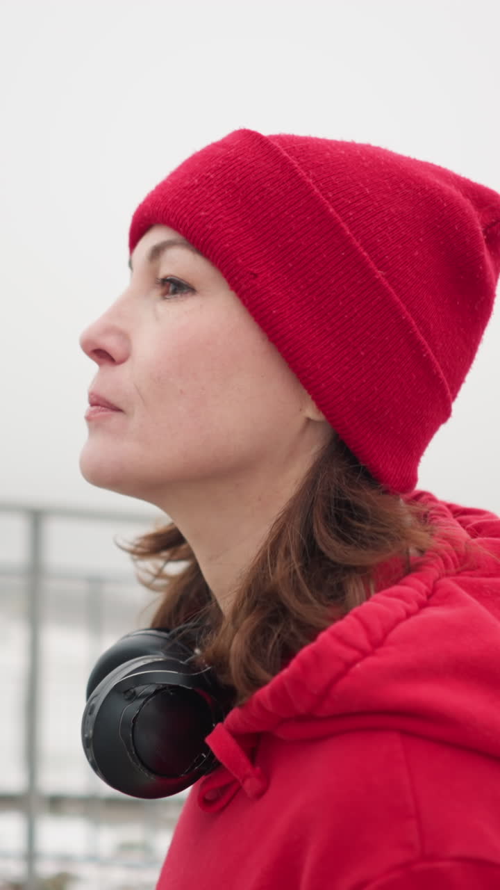 Close-up of woman walking near iron railing wearing red beanie and jacket with headphones around neck, background shows blurry bridge and settlement in foggy snow covered winter atmosphere