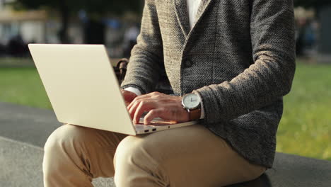 portrait of a good looking businessman sitting on wall in the city park, working on his laptop computer and then smiling at the camera