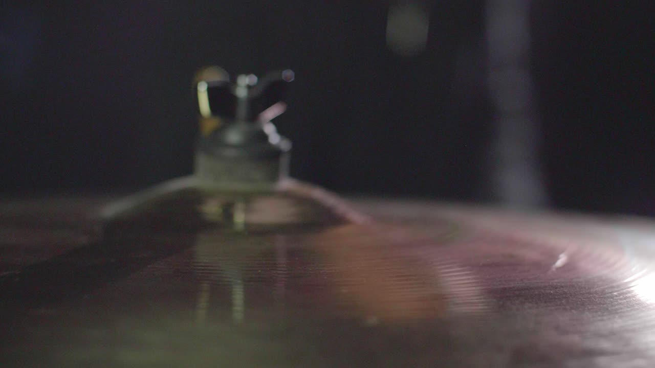 Primo piano fluido di un piatto crash della batteria colpito da una bacchetta. Il piatto oscilla dopo. Sfondo scuro. Super slow motion. Movimento in avanti con dolly gimbal. Profondità di campo ridotta con messa a fuoco fissa.