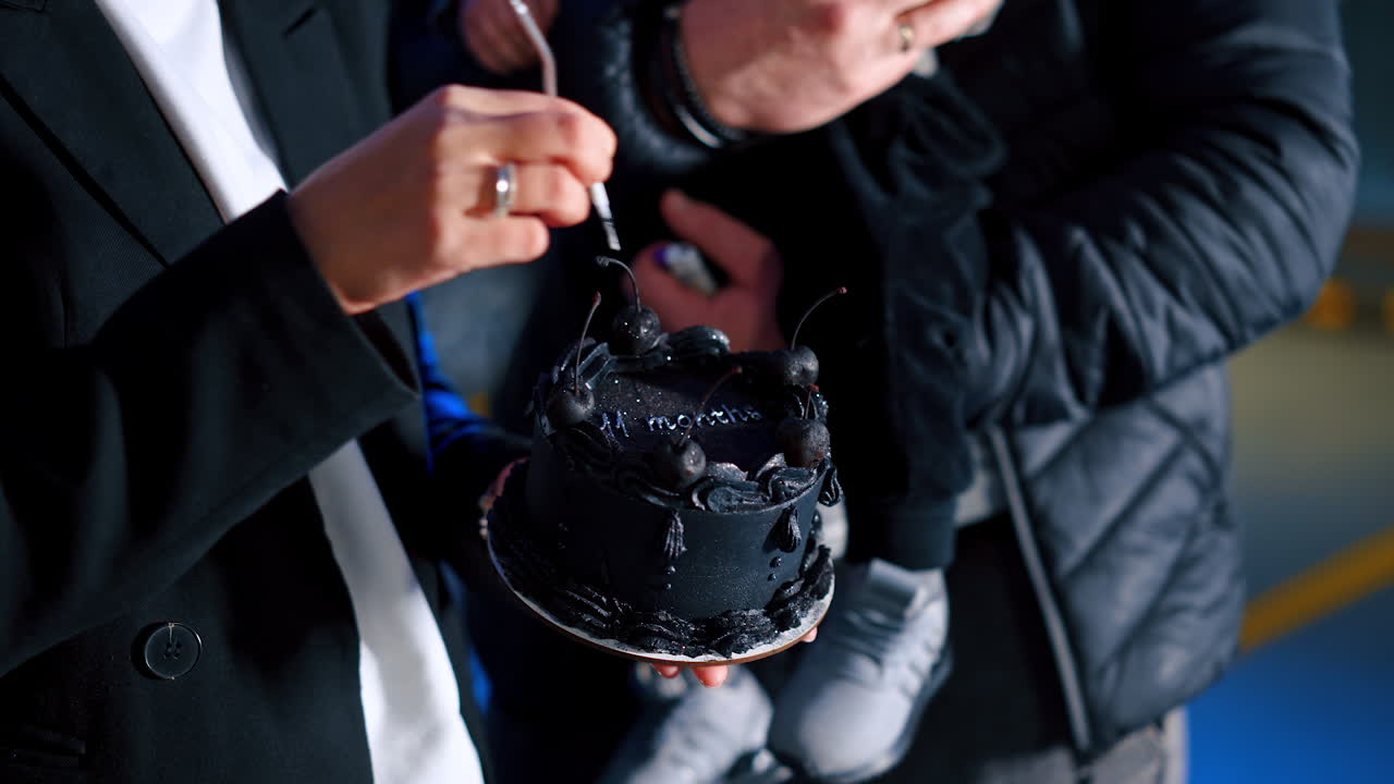 Smiling blonde woman puts a candle on the black cake and lighting it. Man with baby in hands stands watching.