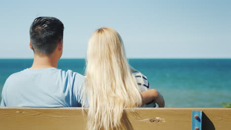 a young couple sits in an embrace on a bench looks at the sea