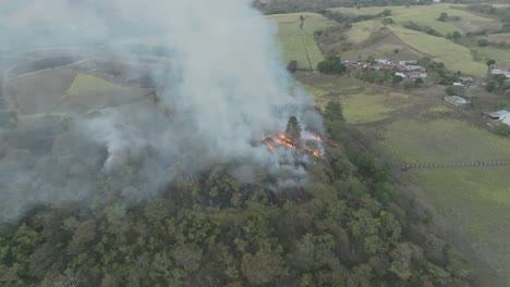 Aerial-drone-shot-in-slow-motion-of-a-mild-forest-fire-on-a-hill-on-a-cloudy-day,-smoke-poised-to-erupt-into-flames