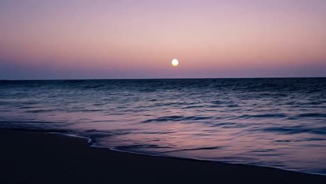 waves crash on a sandy beach as a full moon rises over a purple ocean, creating a serene twilight atmosphere