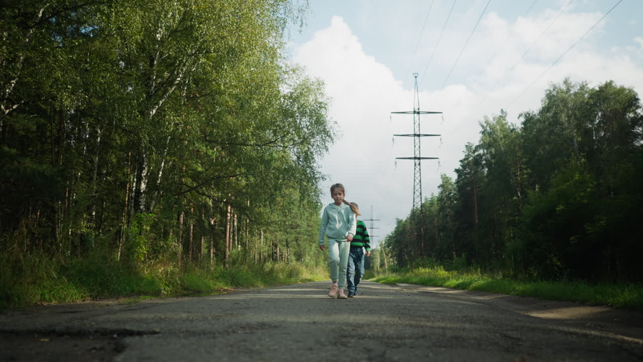 Joyful children walking home along quiet road through forest, swinging hands and smiling in daylight, surrounded by tall trees, green foliage, and overhead power lines under cloudy blue sky