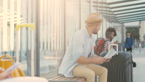 young caucasian handsome man traveller in hat sitting on bench at bus stop and waiting for transport while using a map