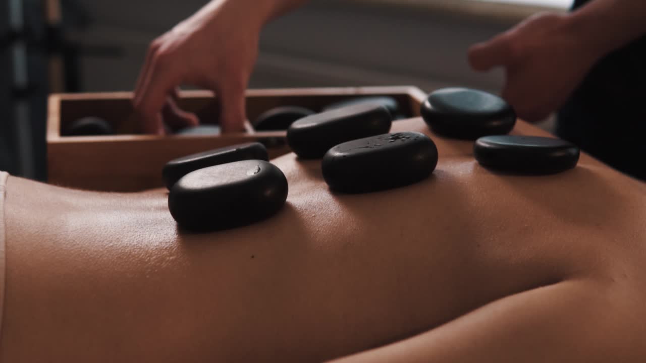 Nærbillede af mandehænder, der placerer varme sorte sten på en smuk kvindes ryg til en særlig opvarmet massage.
