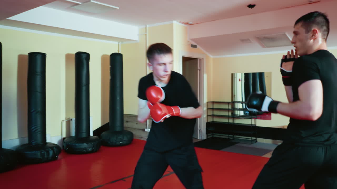 Wrestlers engaged in sparring session on red mat inside training gym, wearing gloves and black outfits, one athlete bending down defending against attack from opponent in active martial arts combat