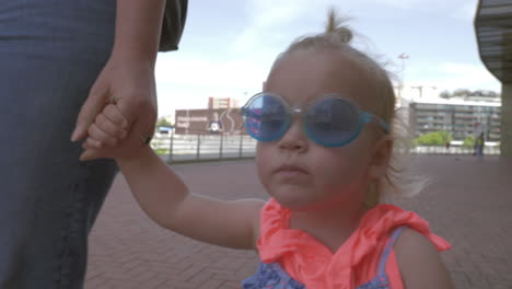 Una-Niña-De-Pelo-Rubio-Con-Unas-Gafas-De-Sol-Redondas-Azules-Muy-Elegantes-Caminando-De-La-Mano-De-Su-Madre