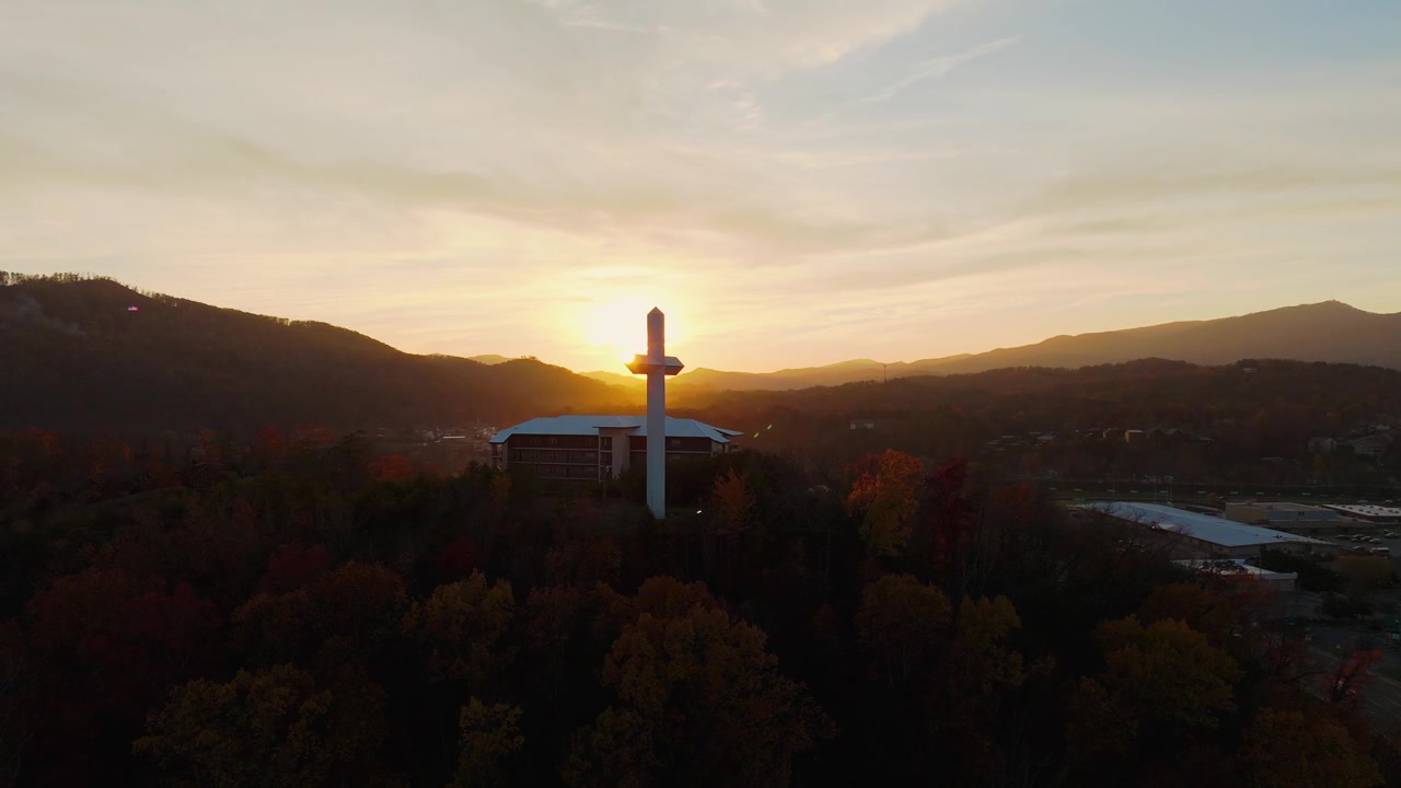 Rekaman drone 4K gerak lambat mengelilingi salib putih menjulang tinggi di atas pepohonan di Tennessee, saat jam emas. Kilatan matahari yang menakjubkan bersinar menembus siluet salib dari berbagai sudut