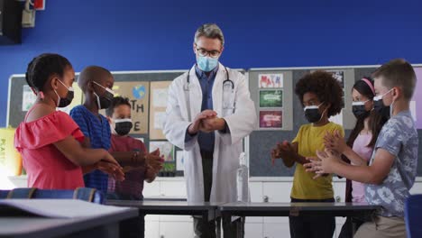 Diverse-schoolteacher-and-schoolchildren-standing-disinfecting-hands,-all-wearing-face-masks