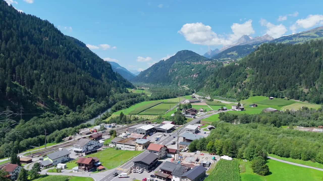 Cars leaving street of swiss town near railway tracks in summer. Aeria wide shot,. Sunny day with blue sky. Green mountains and alps in background