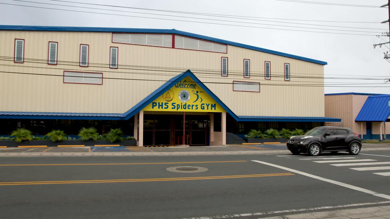 A static wide shot captures cars driving past a school sports facility in Palau