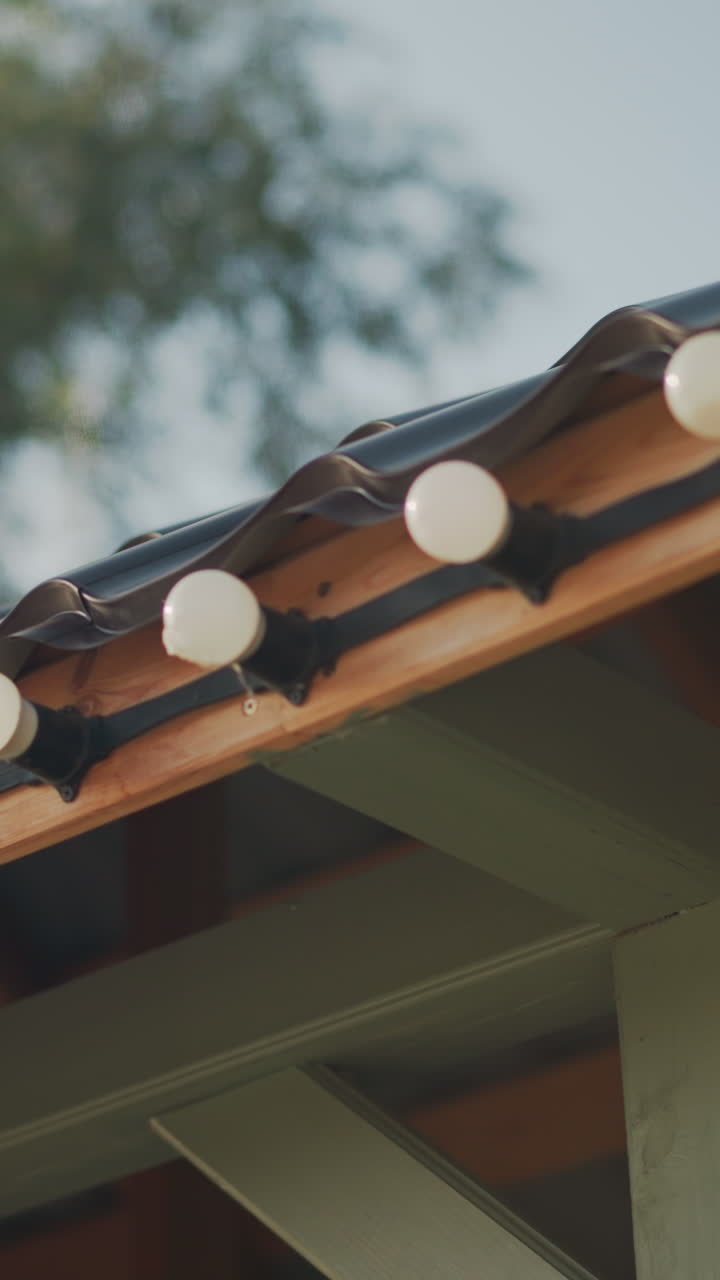 bombillas redondas iluminadas por el sol sobre tejado de tejas, primer plano de luces de fiesta a lo largo del alero de tejas de cerámica, vigas pintadas, cálido resplandor diurno, preparación del patio por parte del propietario, organizador de eventos que organiza la configuración, estilista