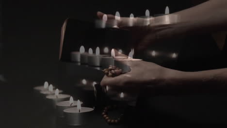 animation of burning candles over hands holding bible and rosary on black background