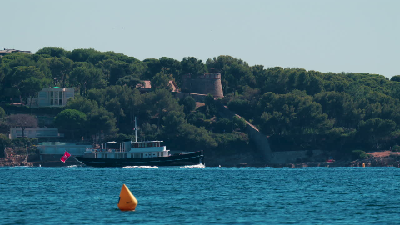 A stylish black yacht sails along the Cannes coastline surrounded by pine trees and villas under clear blue skies on the French Riviera