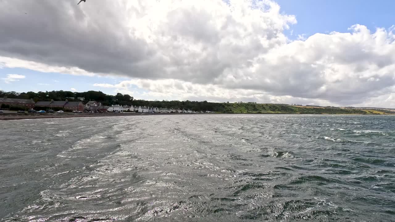 Wide-angle daytime shot pans slowly across Cromarty’s waterfront, capturing choppy water, historic seaside houses, dramatic clouds, and natural coastal scenery