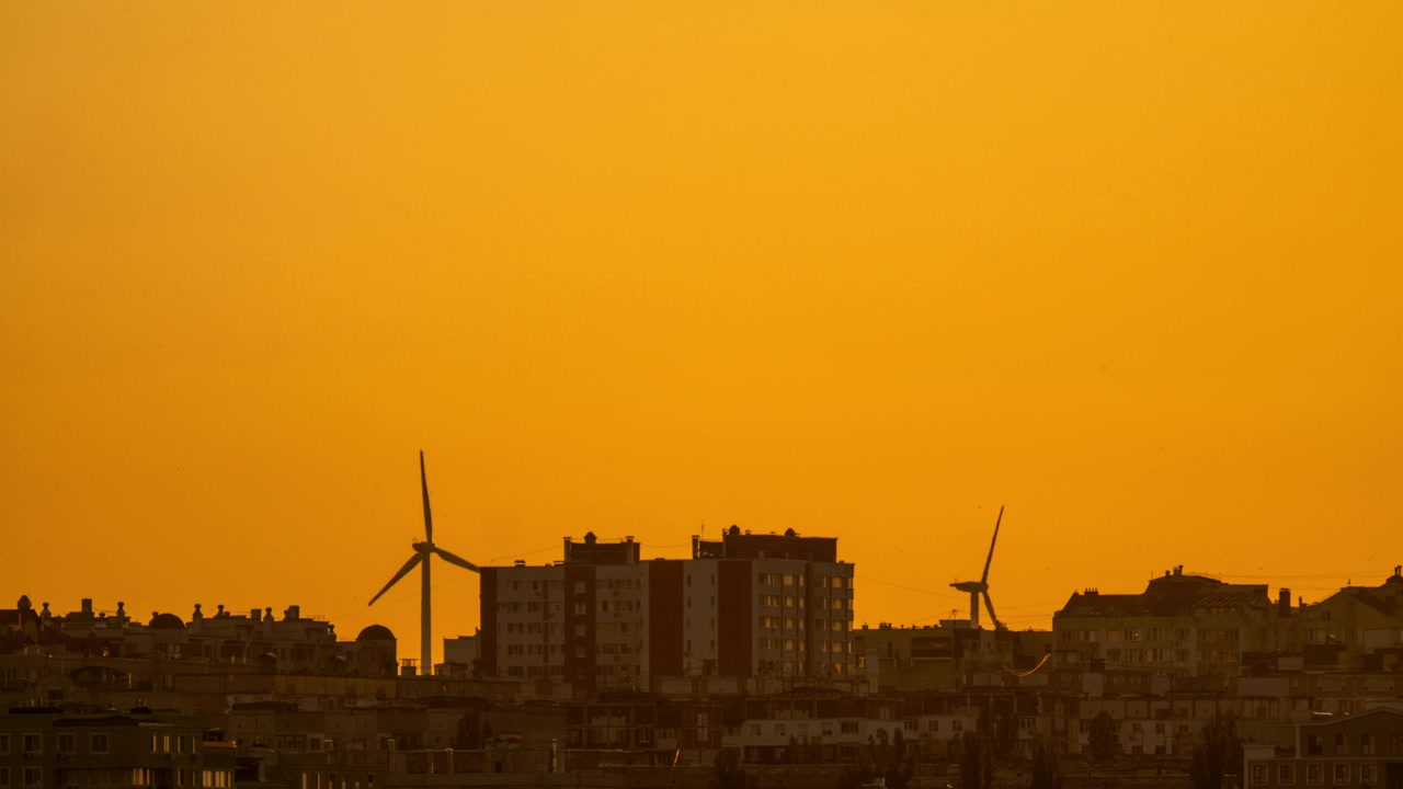 Lapso de tiempo de aerogeneradores silueteados girando sobre el horizonte de una ciudad durante un atardecer dorado