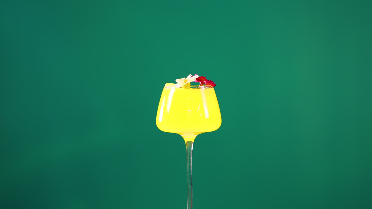 A vibrant yellow cocktail with fruit garnish against a green screen backdrop. Bright lighting highlights the drink's vivid colors