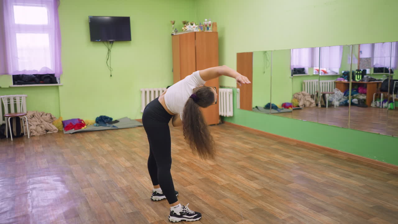 Lady in black leggings and white top stretching her body in front of mirror inside gym studio, raising one arm overhead with reflection visible on large wall mirror and wooden floor background