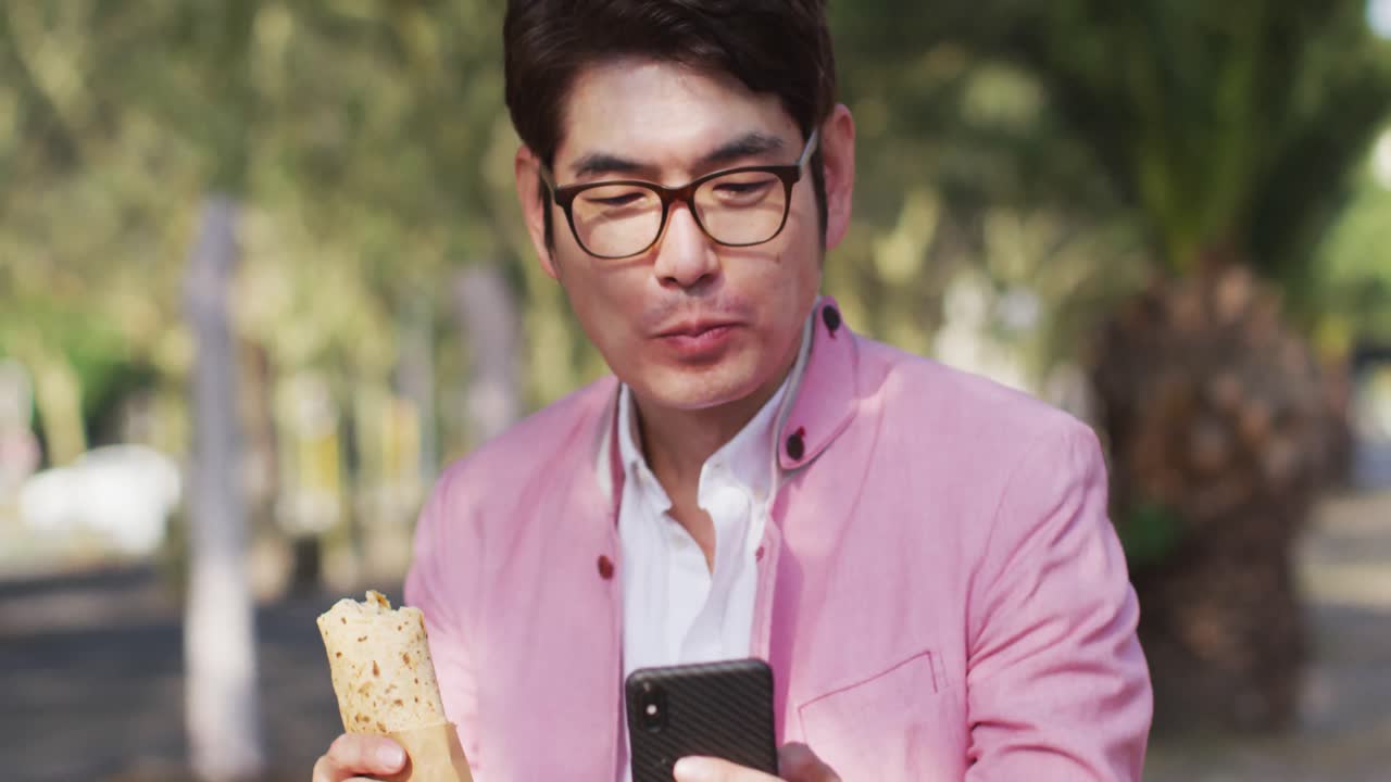 공원 에 앉아 있는 아시아 사람 이 간식 을 먹고 스마트폰 을 사용 하고 있다