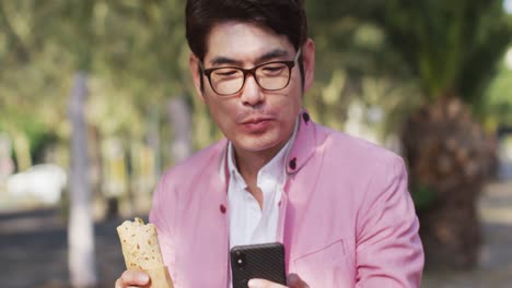 hombre asiático tomando un bocadillo y usando un teléfono inteligente mientras está sentado en el parque