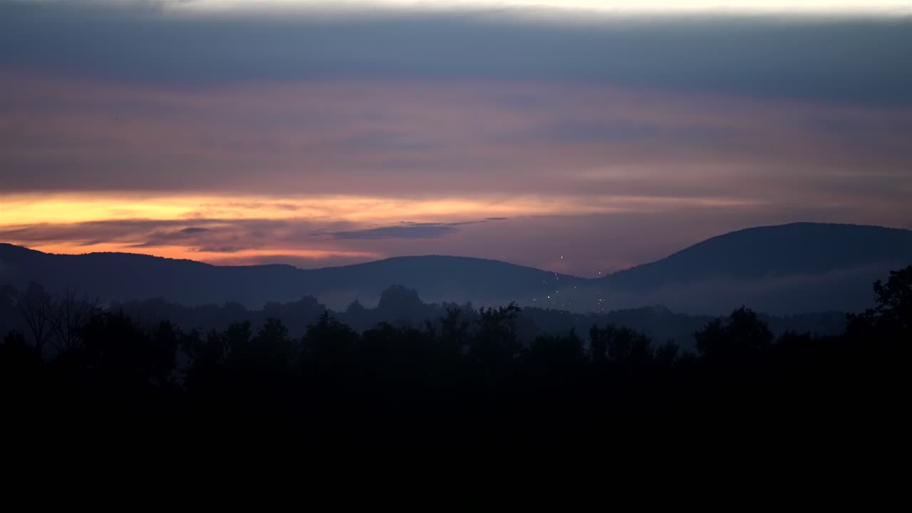 los fuegos artificiales estallan y brillan bajo el resplandor del anochecer del cielo y las montañas frente al bosque