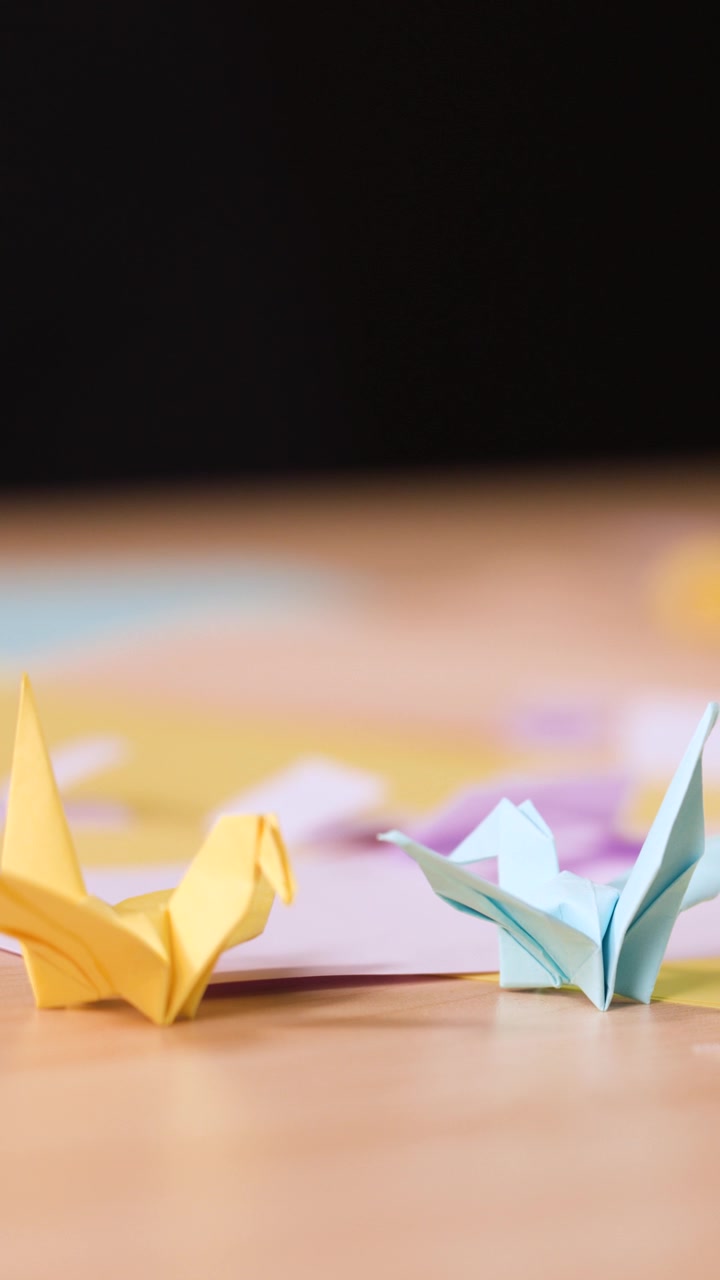 Una persona piega una gru di carta viola accanto a uccelli origami gialli e blu su legno.