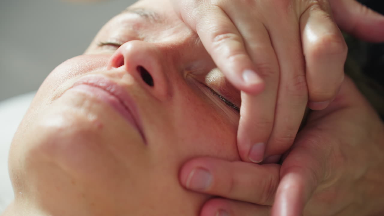Close up view of recipient lying face up on massage bed as beauty expert hands gently smooth facial skin in soft circular motions under soothing spa light for deep relaxation and rejuvenation
