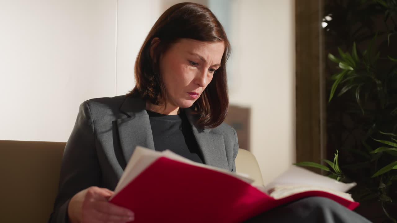 Focused businesswoman reading red folder with serious expression while sitting in office lounge, reviewing important documents or preparing for meeting in professional work environment