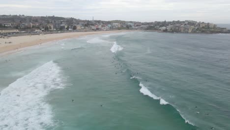 Salpicando-Olas-Con-Turistas-En-La-Playa-De-Bondi-Con-Paisaje-Urbano-De-Fondo-En-Los-Suburbios-Del-Este,-Sydney,-Nueva-Gales-Del-Sur,-Australia