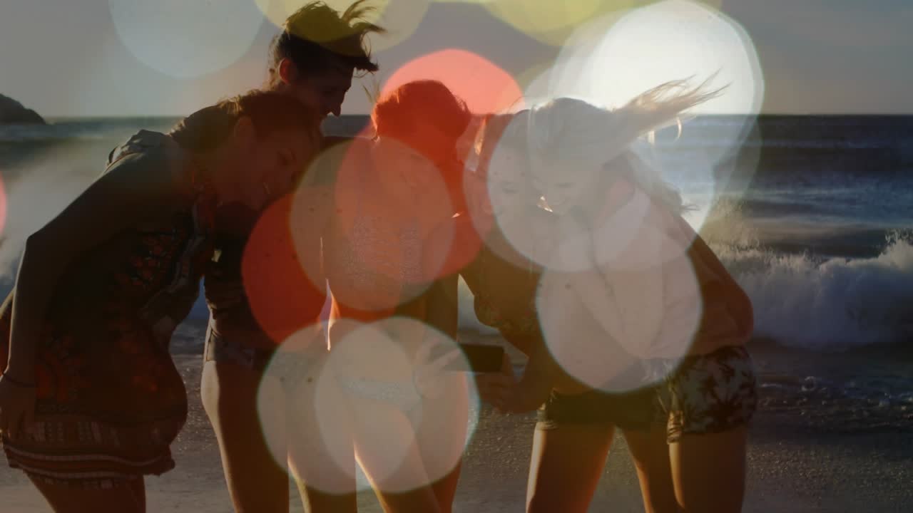Five women huddling on shoreline in swimsuits and cap, one holding phone, others leaning to view