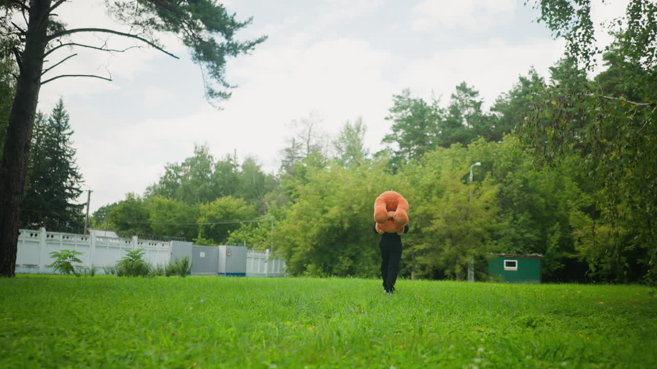 Rear view of kid walking across green field with teddy bear on head, surrounded by tall grass, trees, distant buildings, and a fence, evoking carefree childhood moment in natural outdoor park setting